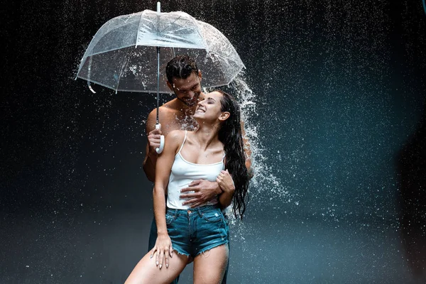 Homem barbudo feliz segurando guarda-chuva e de pé com namorada alegre no preto — Fotografia de Stock