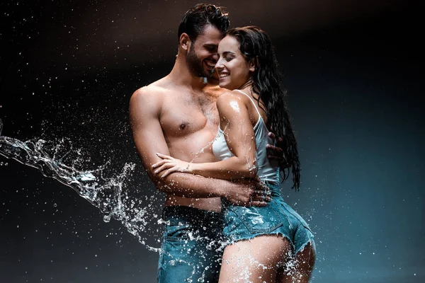 Sin camisa y sexy hombre abrazando feliz novia en pantalones cortos de mezclilla cerca de salpicadura de agua en negro - foto de stock