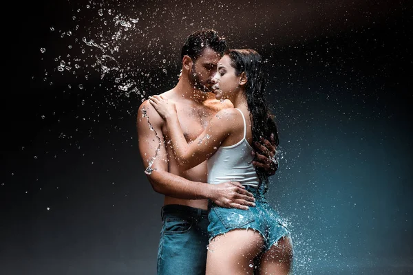 Без рубашки и сексуальный мужчина обнимает подружку в джинсовых шортах возле брызг воды на черном — стоковое фото
