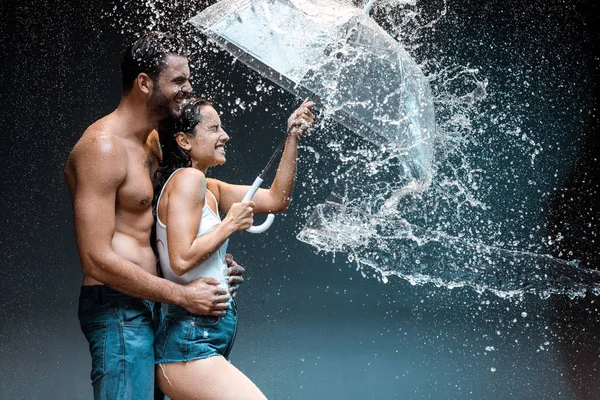 Glücklich bärtiger Mann umarmt fröhliche Freundin mit Regenschirm in der Nähe Spritzwasser auf schwarz — Stockfoto