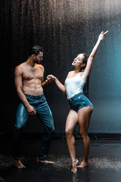 Cheerful couple standing and holding hands under raindrops on black — Stock Photo