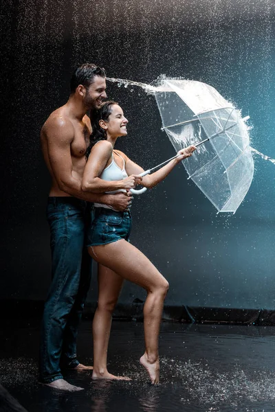 Feliz barbudo homem abraçando atraente namorada segurando guarda-chuva perto respingo de água no preto — Fotografia de Stock