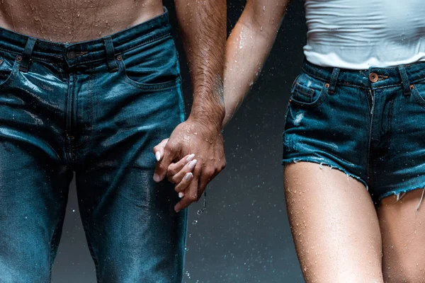 Обрезанный вид мокрого мужчины, держащегося за руки с девушкой в джинсовых шортах на черном — стоковое фото
