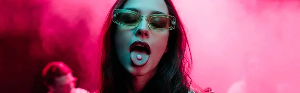 Panoramaaufnahme eines schönen Mädchens mit Sonnenbrille und LSD auf der Zunge in einem Nachtclub mit rosa Rauch — Stockfoto