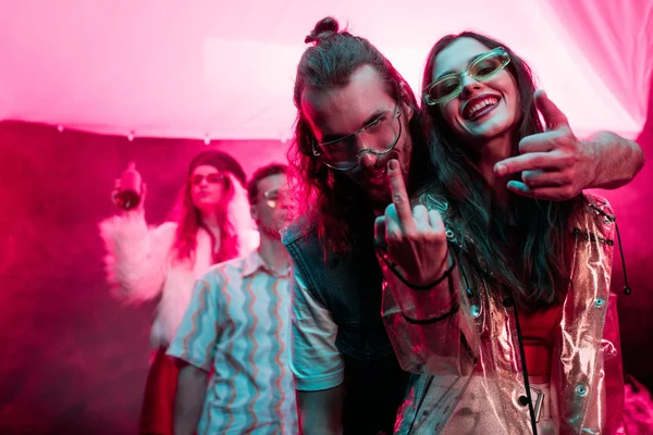Feliz chica y hombre en gafas de sol mostrando los dedos medios en discoteca durante fiesta rave - foto de stock