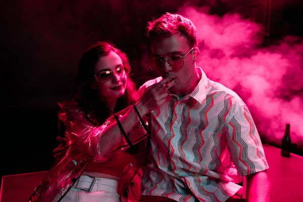 Man smoking cigarette near young woman during rave party in nightclub — Stock Photo