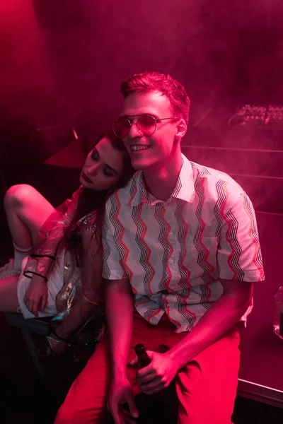 Sonriente hombre con cerveza sentado cerca de chica en discoteca durante rave - foto de stock