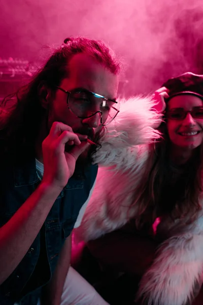 Мужчина курит марихуану рядом с улыбающейся девушкой в ночном клубе — стоковое фото