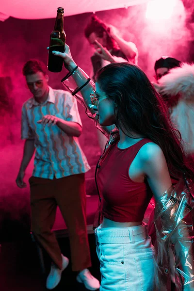Hermosa chica sosteniendo cerveza y bailando en discoteca con humo rosa neón - foto de stock