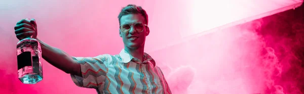 Panoramic shot of handsome man with bottle of alcohol during rave in nightclub — Stock Photo