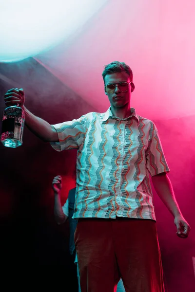 Homme en lunettes de soleil avec bouteille d'alcool pendant la rave dans la boîte de nuit — Photo de stock