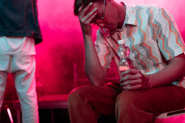 Enfermo con dolor de cabeza sentado con botella de agua en el club nocturno - foto de stock