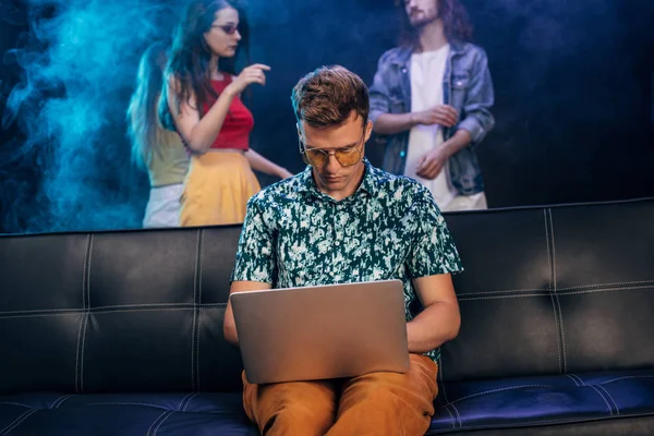 Hombre guapo en gafas de sol sentado en el sofá y el uso de ordenador portátil en el club nocturno - foto de stock