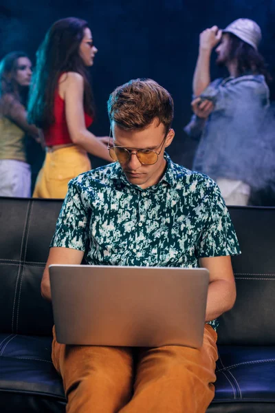 Schöner Mann mit Sonnenbrille sitzt auf Sofa und benutzt Laptop in Nachtclub — Stockfoto