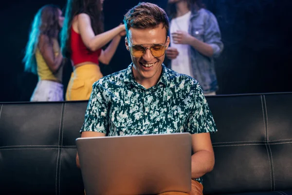 Man in sunglasses sitting on couch and using laptop in nightclub — Stock Photo