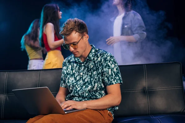 Man in sunglasses sitting on couch and using laptop in nightclub — Stock Photo