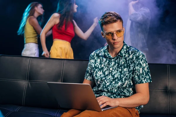 Man in sunglasses sitting on couch and using laptop in nightclub — Stock Photo