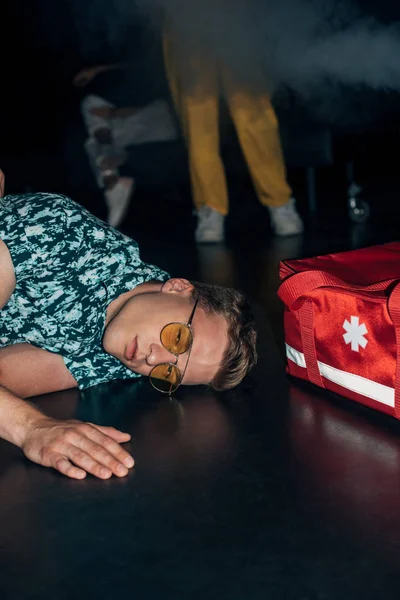 Homme en lunettes de soleil couché sur le sol près du sac trousse de premiers soins dans la boîte de nuit — Photo de stock