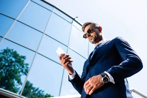 Vista de ángulo bajo del hombre guapo en traje y gafas usando teléfono inteligente - foto de stock