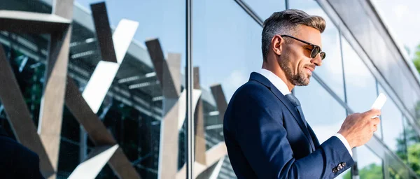 Plano panorámico del hombre guapo en traje y gafas usando teléfono inteligente - foto de stock