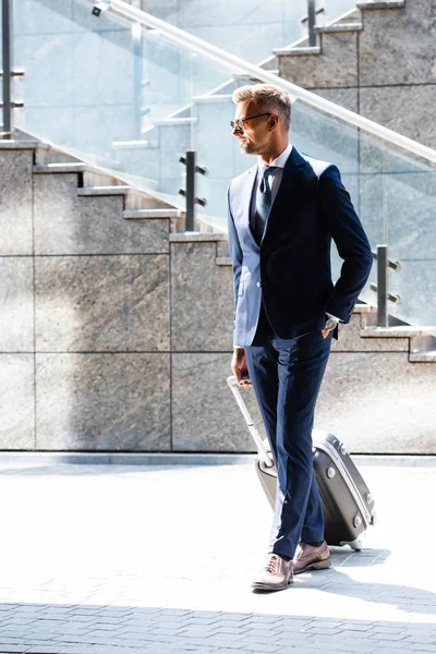 Hombre guapo en traje y gafas con la mano en el bolsillo que sostiene la maleta - foto de stock
