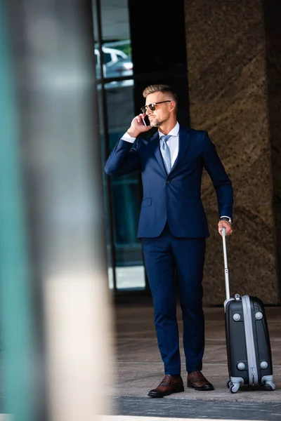 Homem de negócios bonito em terno e óculos falando no smartphone e segurando mala — Fotografia de Stock