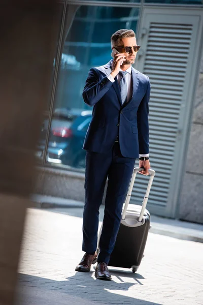 Foco seletivo de empresário bonito em terno e óculos falando no smartphone e segurando mala — Fotografia de Stock
