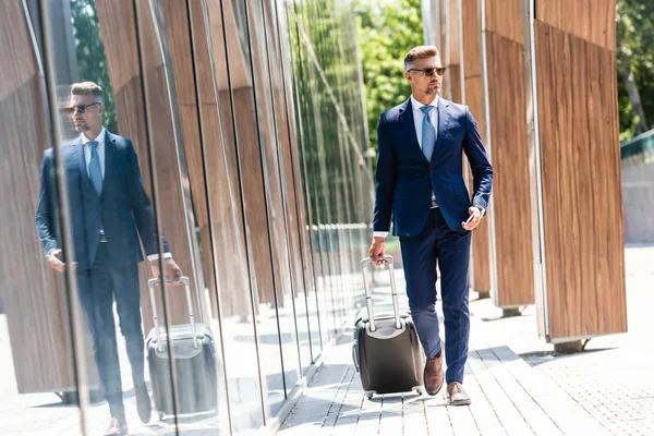 Bel homme d'affaires en costume et lunettes tenant smartphone et valise — Photo de stock