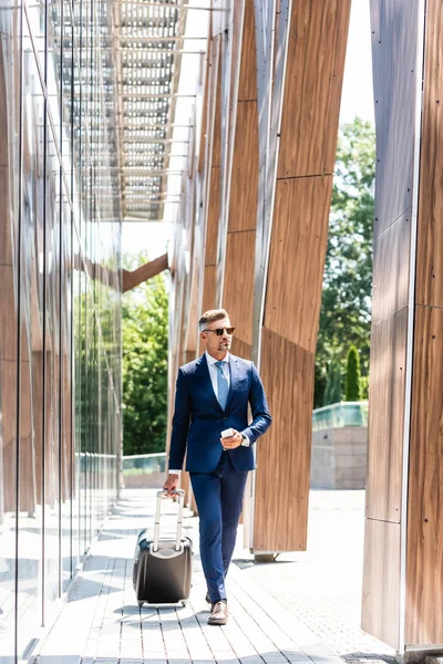Hombre de negocios guapo en traje y gafas que sostienen el teléfono inteligente y la maleta - foto de stock