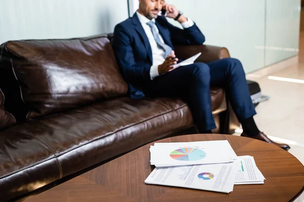 Enfoque selectivo de los papeles en la mesa y el hombre hablando en el teléfono inteligente en segundo plano - foto de stock
