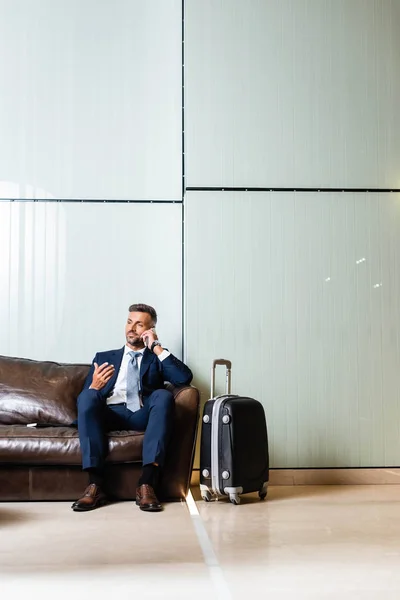 Hombre de negocios guapo en traje sentado en el sofá y hablando en el teléfono inteligente - foto de stock