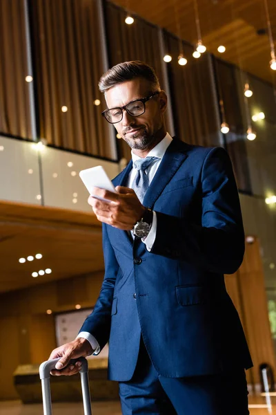 Bel homme d'affaires en costume et lunettes en utilisant smartphone à l'hôtel — Photo de stock