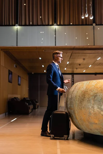 Vue latérale du bel homme d'affaires en costume et lunettes tenant la valise — Photo de stock