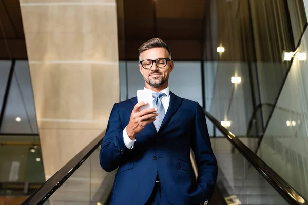 Hombre de negocios guapo en traje y gafas usando teléfono inteligente en el hotel - foto de stock