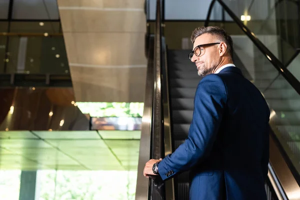 Back view of handsome businessman in suit and glasses on escalator — Stock Photo