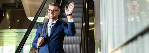 Panoramic shot of handsome businessman in suit and glasses waving in hotel — Stock Photo