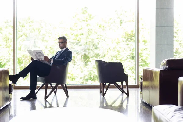 Handsome businessman in suit sitting on armchair and reading newspaper — Stock Photo