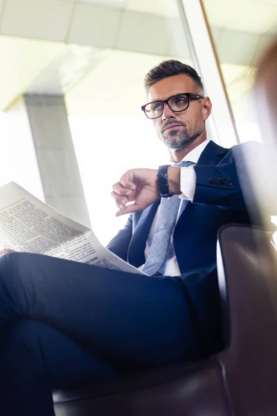 Guapo hombre de negocios con traje y gafas mirando hacia otro lado y sosteniendo el periódico - foto de stock