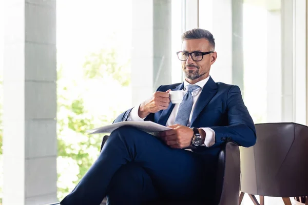 Handsome businessman in suit and glasses holding cup and newspaper — Stock Photo