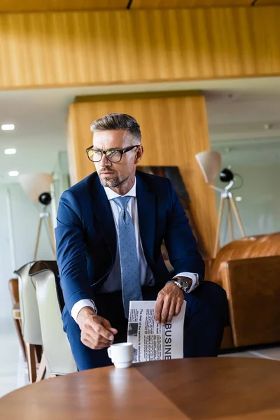 Красивий бізнесмен в костюмі і окулярах бере чашку і тримає газету — стокове фото