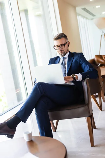 Homem de negócios bonito em terno e óculos com fones de ouvido usando laptop — Fotografia de Stock