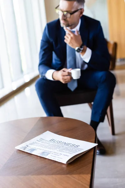 Selective focus of newspaper on table and handsome businessman in suit with cup on background — Stock Photo