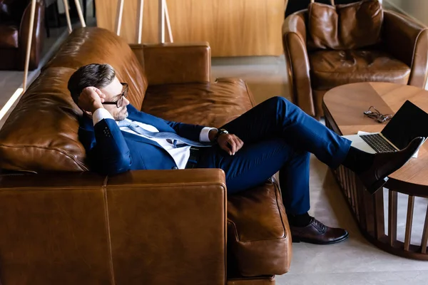 Side view of handsome businessman in suit and glasses sitting on sofa — Stock Photo