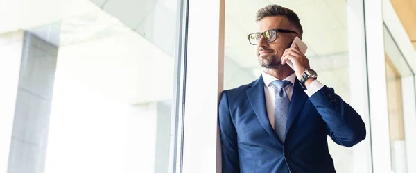 Plano panorámico de hombre de negocios guapo en traje y gafas hablando en smartphone - foto de stock