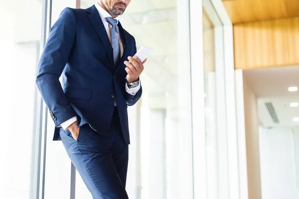 Vista recortada del hombre de negocios en traje con la mano en el bolsillo utilizando el teléfono inteligente - foto de stock