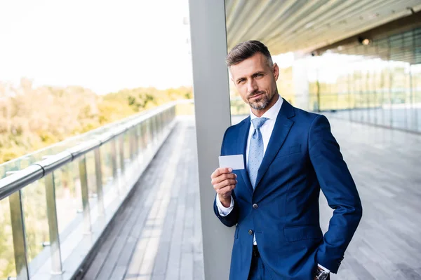 Homem de negócios bonito em desgaste formal com a mão no bolso segurando cartão em branco — Fotografia de Stock