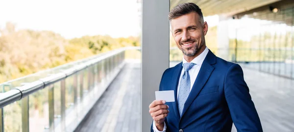 Panoramic shot of handsome businessman in formal wear holding blank card — Stock Photo