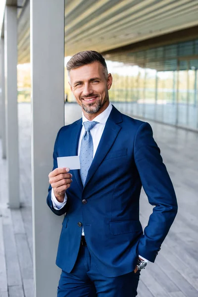 Guapo hombre de negocios en ropa formal con la mano en el bolsillo celebración de la tarjeta en blanco - foto de stock