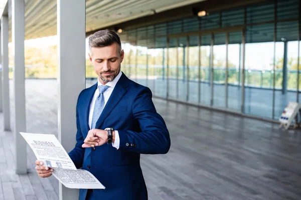 Handsome businessman in formal wear holding newspaper and looking at watch — Stock Photo