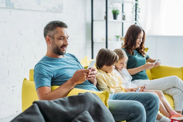 Happy parents with daughter and son using smartphones while sitting on sofa at home — Stock Photo
